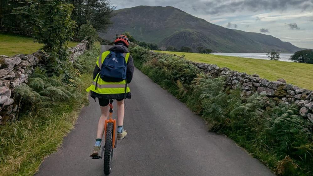 A person pedalling away from the camera on a unicycle along a pathway, with mountains in the background.
