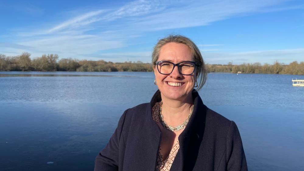 Helen Harrison with light-coloured hair tied back and large blue glasses wearing a dark coat and standing next to a large lake surrounded by trees