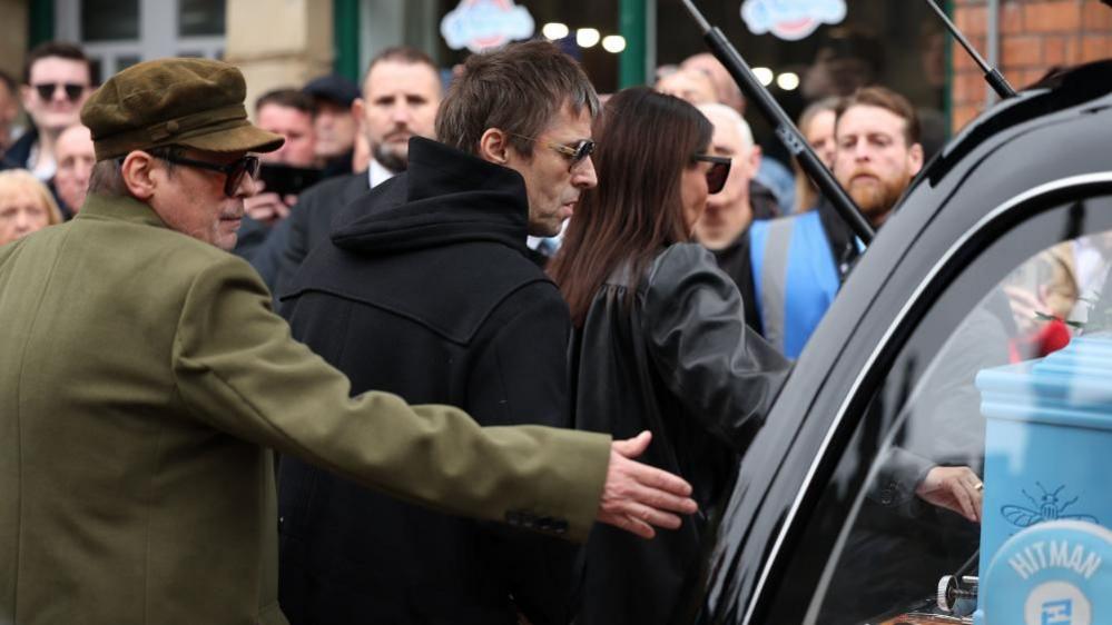 Liam Gallagher stands wearing a black jacket and sunglasses looking at a hearse with a blue coffin inside.