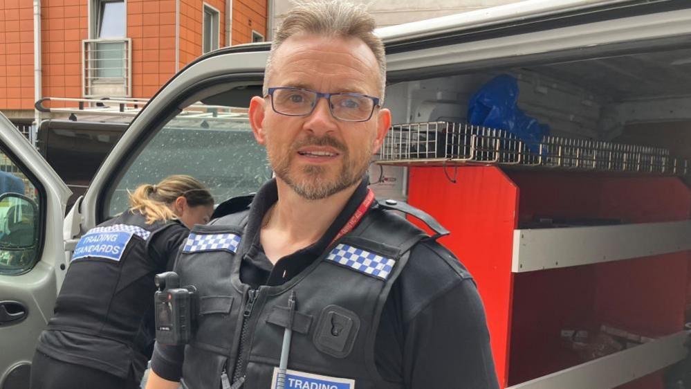 Stuart Hughes standings in front of a van where he is bagging evidence. His colleague is in the background to the left of the picture, leaning into the van's front passenger seat. Both are wering black polo shirts with stab vests over the top. There is a bodyworn camera on Mr Hughes' chest. He is wearing glasses. His vest says "TRADING STANDARDS" in white letters on a blue badge.