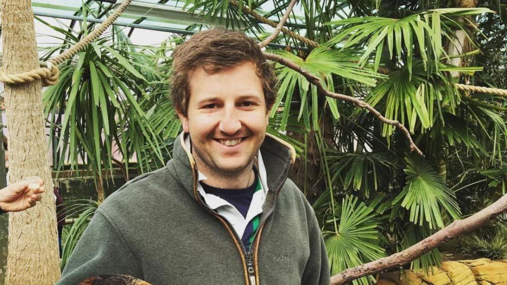 Max Hunter has short brown hair and is smiling. He is wearing a green jumper and is standing in front of a large green plant.