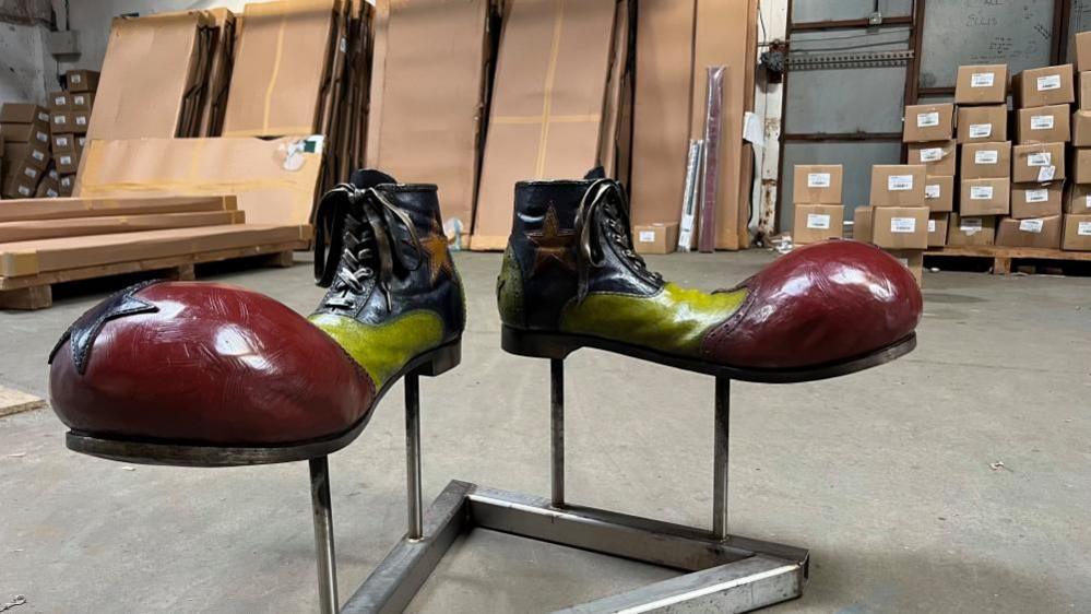 Two giant clown shoes, painted in black uppers, with yellow mid banding, and red enlarged toecaps, stands on a stainless steel frame that will be buried underground. In the backdrop is a series cardboard boxes of various size.