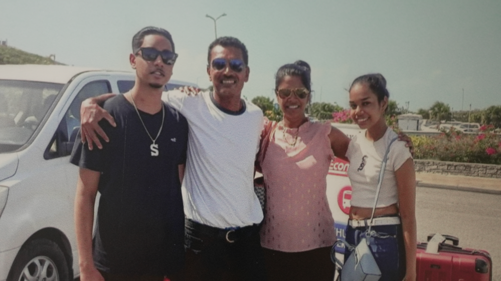 Shawn Seesahai with his parents and sister; they all smile at the camera with their arms around each other. They are stood outside and there is a vehicle behind them to their left, and a suitcase is to their right