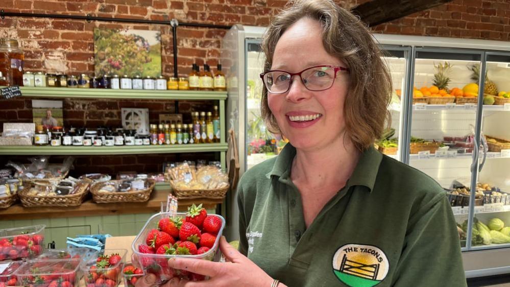 Emma Tacon stands in her farm shop, wearing a green polo shirt, branded with her farm shop logo. Emma has long straw-blonde hair. She is holding a punnet of strawberries. Behind her are display fridges, shelves and worktops stocked with local produce.