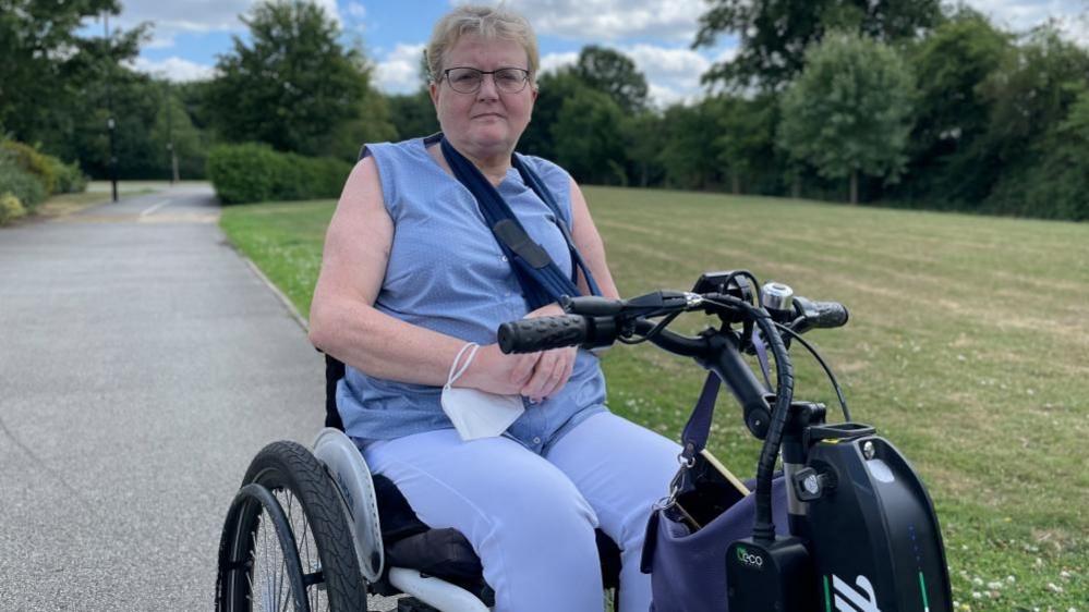 Disability activist Flick Williams pictured in a park, wearing a blue sleeveless top and blue trousers. She sits in a wheeled motorised vehicle. 