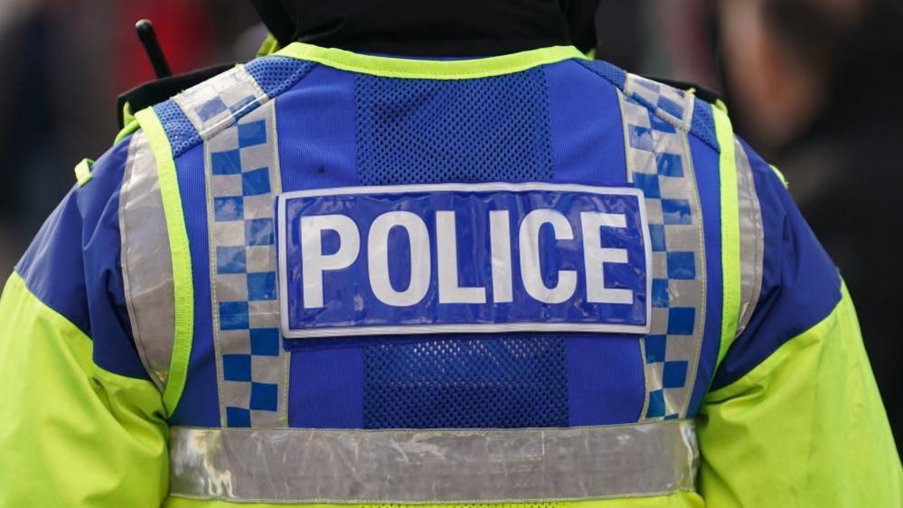 A police officer with his back to the camera wears a yellow and blue high-vis jacket with the word 'police' on the back. 