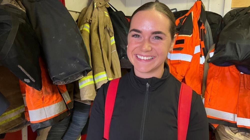 A woman with hair tied back looks into the camera and smiles. She is wearing a black top with red braces holding up firefighting uniform trousers. She stands in front of firefighters' uniforms hanging on pegs along a wall.
