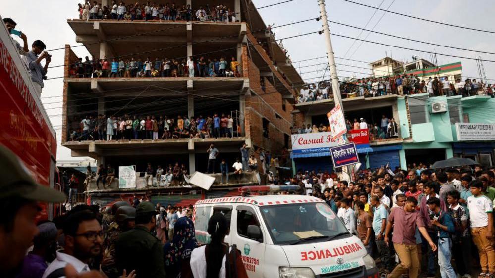 Crowds of people stand on the streets and on unfinished buildings looking down at an ambulance which makes its way through the crowd.