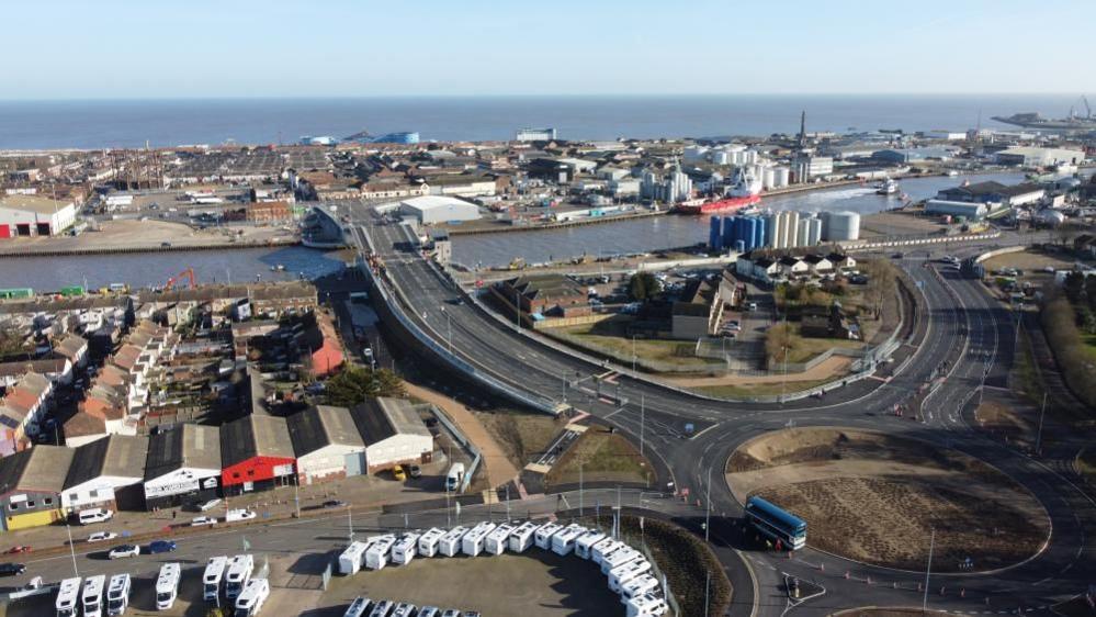 Herring Bridge and the associated four lane road approaches from the Southtown side of the river, looking eastwards to the sea. In the foreground are some motorhomes in a sales yard, with warehousing behind that. Shipping supply tanks and other associated port infrastructure and in the middle distance the Great Yarmouth Pleasure Beach roller coaster is visible.