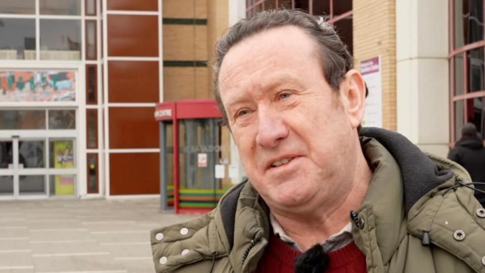 Aidan McGurran standing outside a building with large windows, brown panels and two entrance ways. Mr McGurran has swept back brown hair, and is wearing a khaki jacket and red jersey.