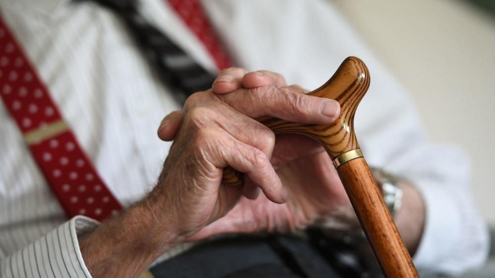 An elderly man sits with a cane in his hand.