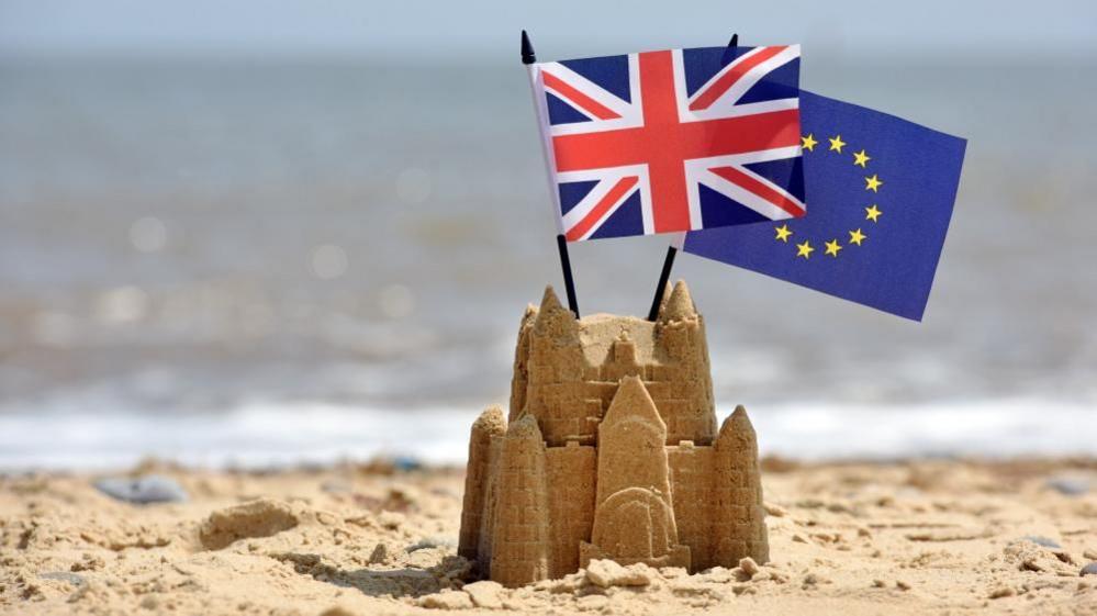 A sandcastle on the beach at Southwold in Suffolk. The North Sea is in the background. It has the Union Jack and European Union flag sticking out of the top of it. 