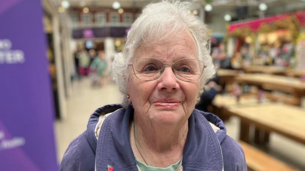 Marjorie Armstrong smiles, wearing a purple hooded coat and jade green top with a poppy badge, with blurred tables in the background at Bolton Market