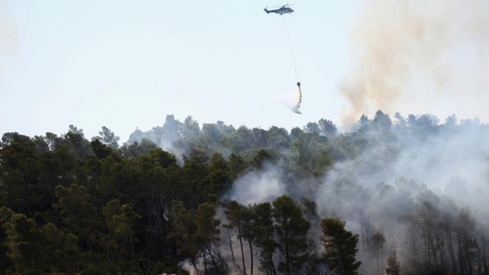 A helicopter battles a wildfire while firefighters work on ground, near Saint-Laurent-de-la-Cabrerisse