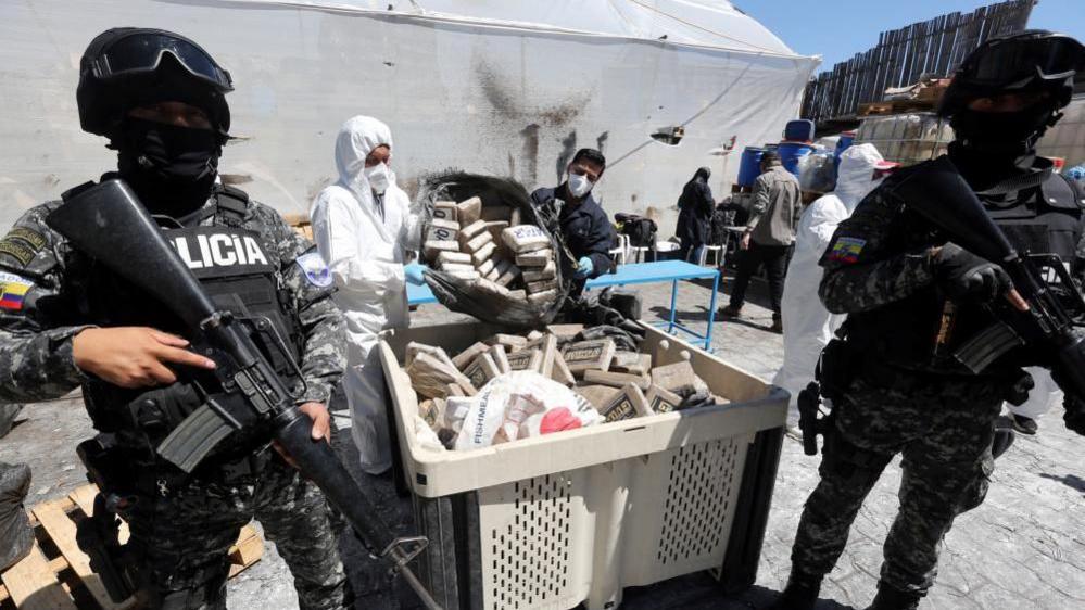 Police with guns stand guard over a container filled with drug packages as two people in masks and hazard clothes tip more drugs inside