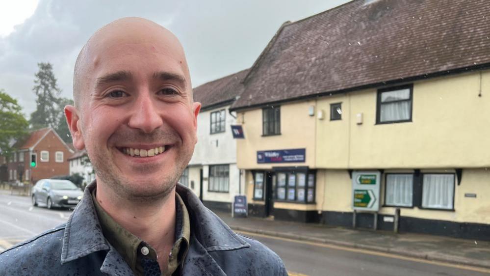 Ben Goldsborough, standing in Long Stratton High Street. He is wearing a khaki shirt, and a blue collared overcoat. He has shaved his head and is smiling. The A140 is behind him, with some painted timber-framed buildings on the opposite side of the road.