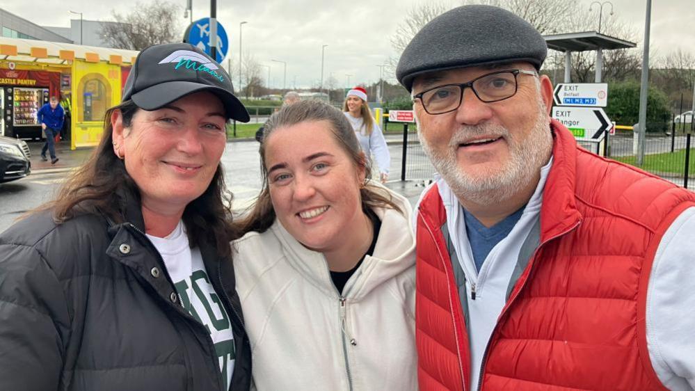 Mary, left, is wearing a black cap and has long brown hair. She is wearing a black coat and white sweatshirt. Caoimhe is wearing a cream hoodie and has brown hair tied back. Mark, right, is wearing a flat cap and thin framed glasses. He has a red body warmer on and a white zip up jumper underneath. 