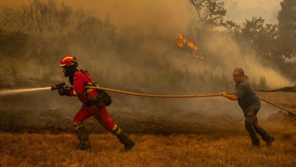A firefighter and a civilian holding a large yellow hose run across a dried out yellow patch of grass with smoke and fires in the woodland behind
