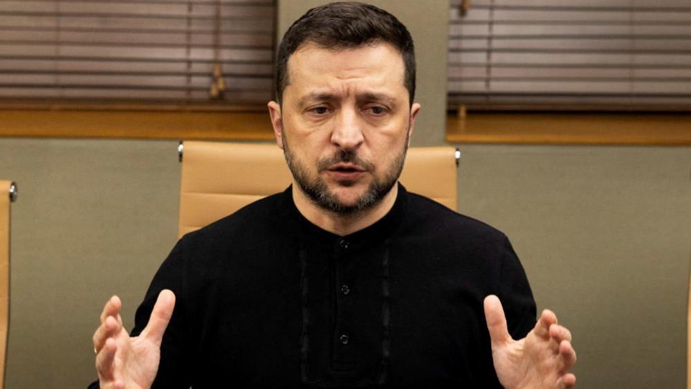 Volodymyr Zelensky sits in a black top with his hands outstretched 