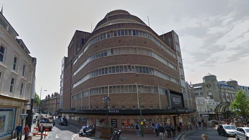 A photo of the building as it was in 2016. It's a seven-story brick building with a curved facade, on the corner of a busy high street with lots of pedestrians and cars