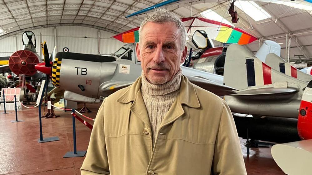 Paul Hennessey, wearing a polo neck jumper and tan winter coat, is looking at the camera. Behind him is a World War 2 plane and museum artefacts. 