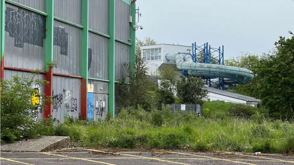 Overgrown grass and bushes next to a derelict looking leisure centre. There is graffiti on the walls of the building and an old dirty water flume can be seen coming out of the swimming pool. 