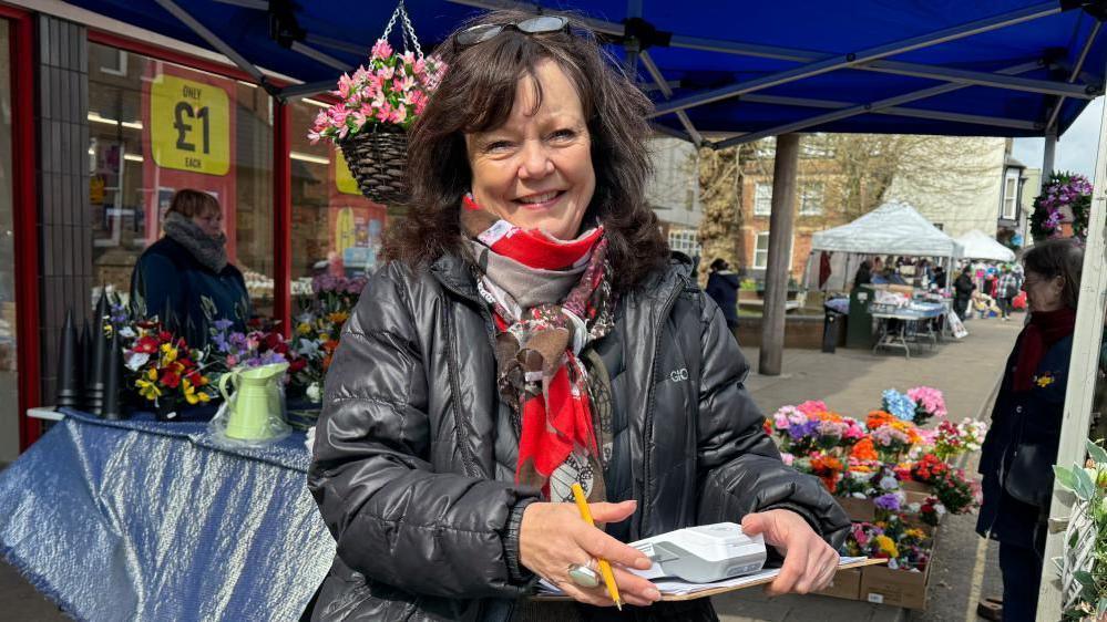 Kate stood in front of a flower stall and smiling, she's wearing a black puffa jacket and a red patterned scarf. She's holding a clipboard and card reader. Behind her you can see a flower stall, sheltered by a blue gazebo.