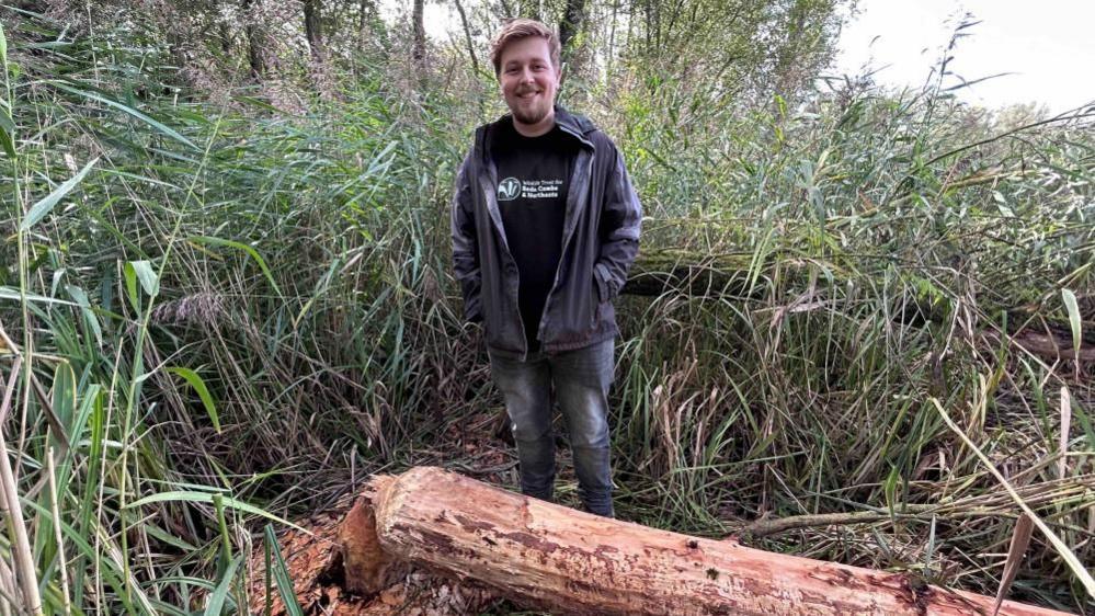 Ben Casey, standing outside, in a nature reserve, but a felled tree, he is looking at the camera and smiling. There is greenery around him. He has short dark hair, a beard, and is wearing jeans, a black T-shirt, a jacket and has his hands in his pocket. 