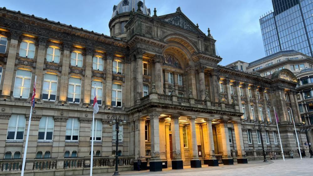 The exterior of the Council House, headquarters of Birmingham City Council. The building has four flagpoles outside it, and comprises of a series of columns and a large archway above the entrance which is lit up by yellow spotlights. 