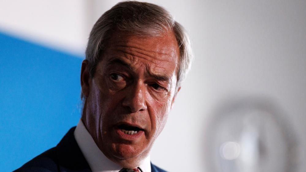 Headshot of Nigel Farage speaking a news conference. He is wearing a dark suit and multi-coloured tie.