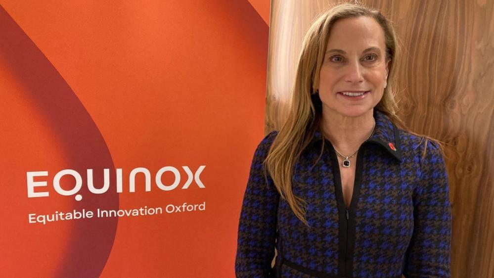 A smartly dressed woman is smiling at the camera. Behind her is a banner with the words EQUINOX, Equitable Innovation Oxford.