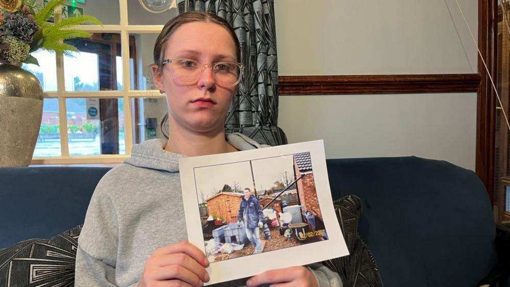 Kayce (pronounced Casey) Burton is sitting on a blue sofa, holding a picture of her dad. He is on a building site, where he had been fitting windows. He is wearing blue jeans and a blue jacket, and there is a shed and tools and the back of a house in the photograph. Kayce is wearing a grey hoodie, and glasses, and has her hair tied back in a ponytail.