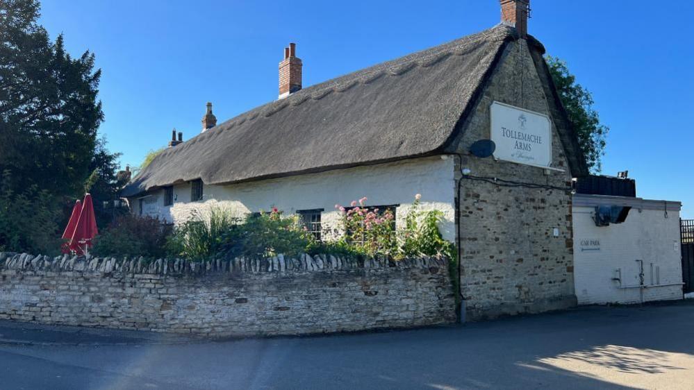 The Tollemache Arms in Harrington with a thatched roof and white walls, with a low stone wall in front of the pub.