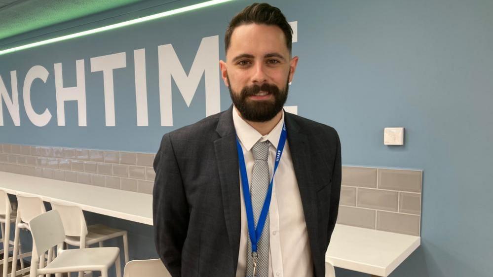 Tom Duffell, a teacher, with dark hair and a beard, wearing a suit jacket, tie, with a lanyard round his neck