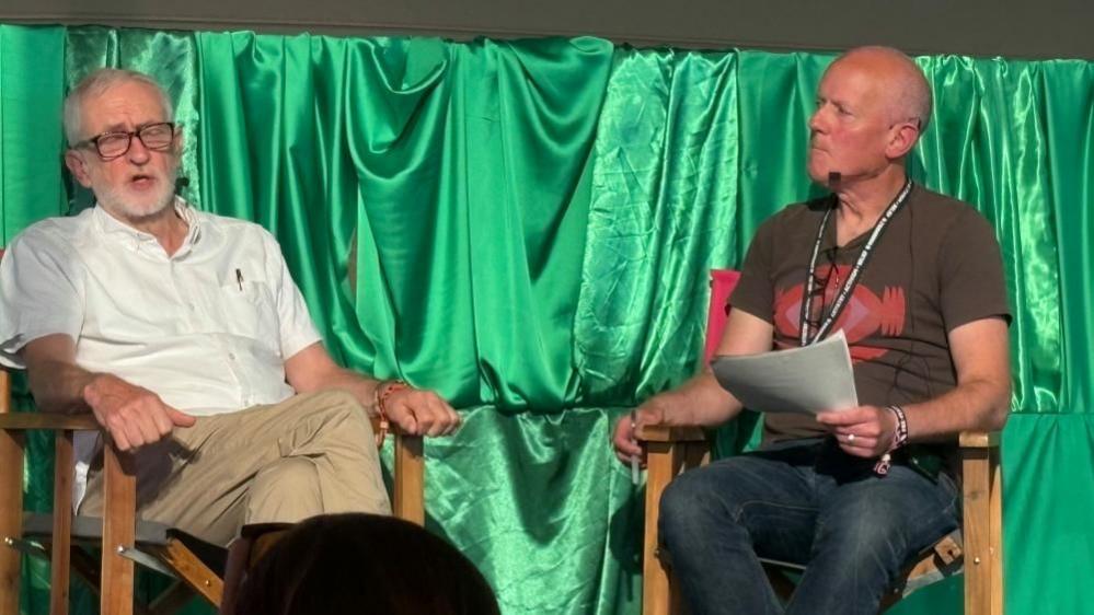 Jeremy Corbyn with short hair and beard, wearing a white short-sleeved shirt, beige trousers and black shoes and Paul Northup, with short white hair, khaki T-shirt, blue jeans and brown shoes, holding a piece of paper. Both are sitting in wooden director-style chairs in front of a green silk curtain on a stage