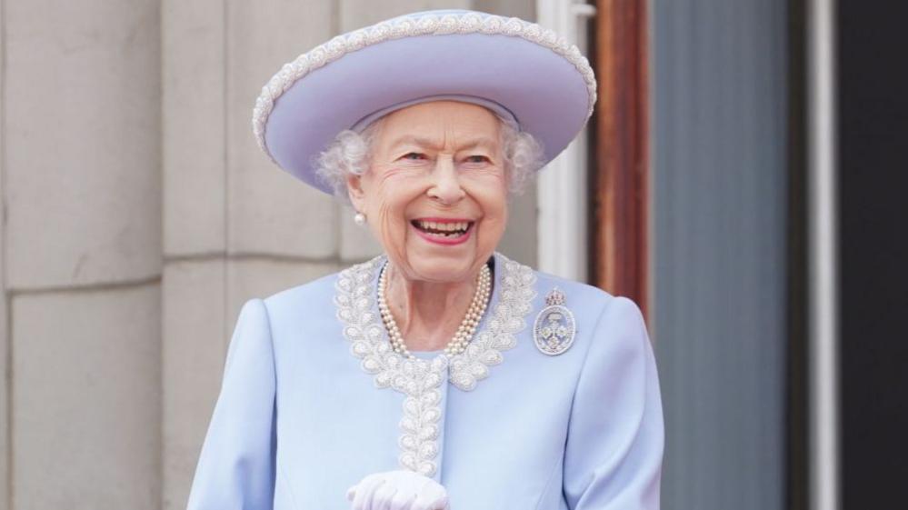 The late Queen Elizabeth II smiles broadly as she stands outside, wearing a lilac hat and jacket - her white gloves can just be seen and she has several strands of pearls around her neck