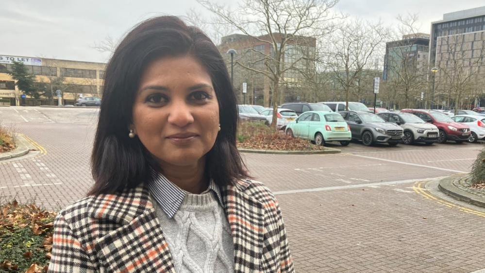 Shazna Muzammil with long dark hair wearing a brown, grey and red patterned jacket and a white top. She is standing in a car park with tall buildings in the background.