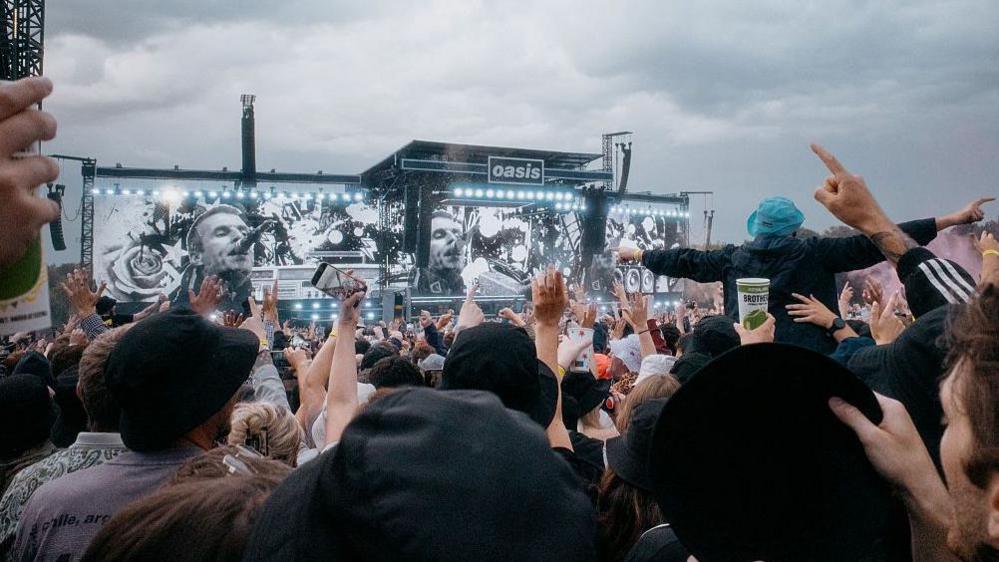 A shot of a large crowd with their backs to the camera. The look on at a big black stage with three identical screens which record a man singing into a microphone with short brown hair. The sign in the centre of the stage reads 'Oasis.' The sky above is grey and cloudy.