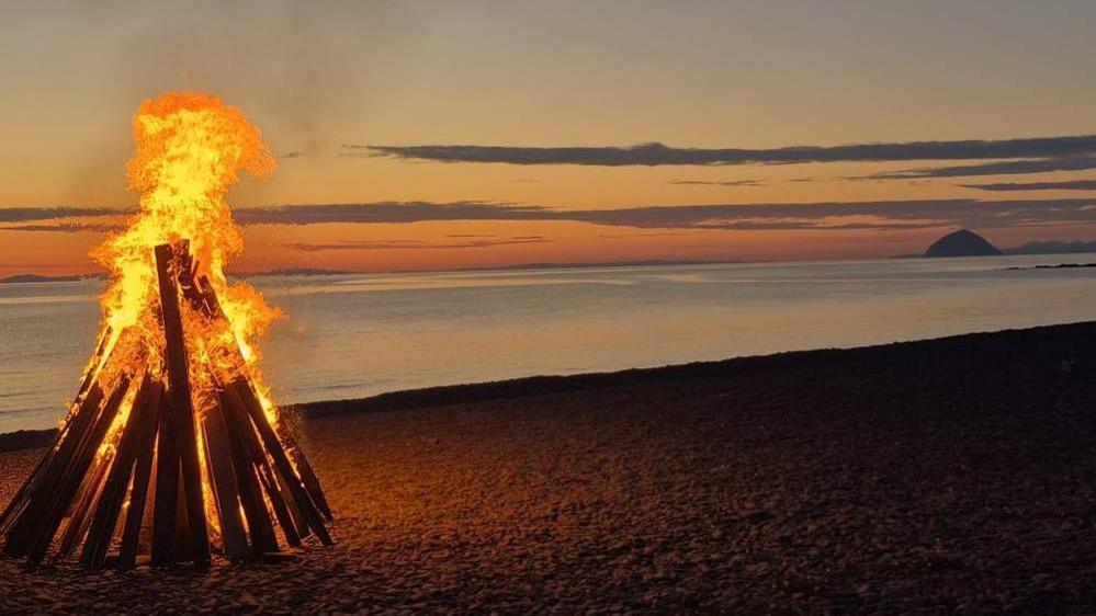Orange flames emerging from a beacon made from wood. It is on a beach in front of a yellow and blue sunset.