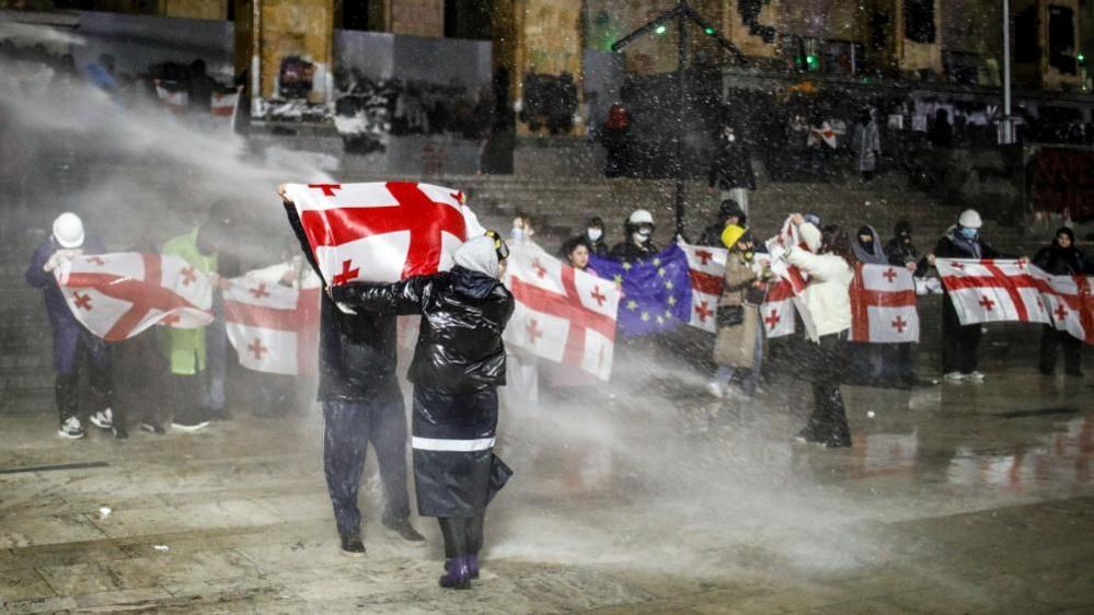 Protesters holding Georgian flags are doused with water cannon outside the Georgian parliament