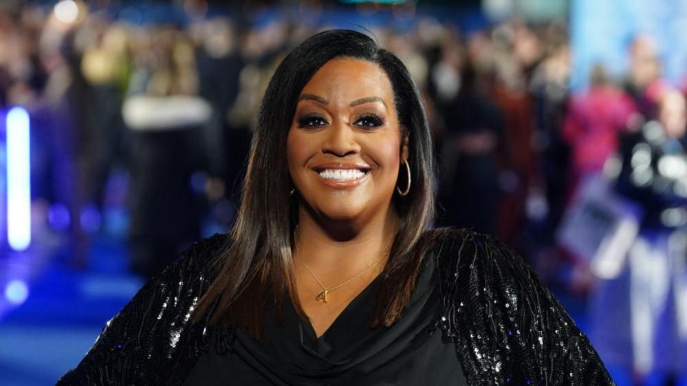 Alison Hammond, smiling towards the camera, wearing a black glittery top, hoop earrings and a gold necklace with an "A" pendent. 