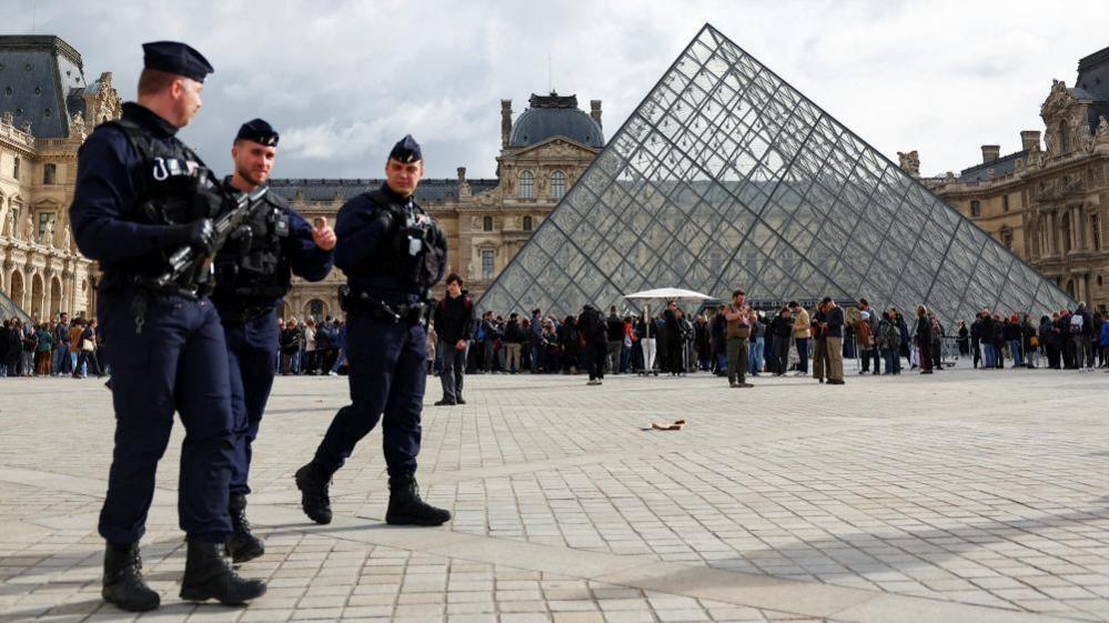 Three French police officers, one carrying a firearm, are seen walking on the square in front of the glass pyramid structure of the Louvre in Paris