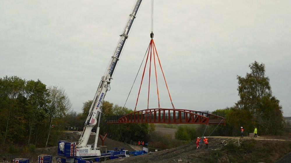 A photograph of the bridge being lowered into place by a large white crane. There are engineers helped the bridge into place. The bridge is surrounded by trees.