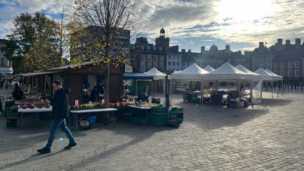 A man walking through a Market Square, showing lots of buildings in the distance, a number of market stalls, white gazebos to the right and wooden ones to the right. There are trees, a water feature in the distance to the right and a fruit and vegetable stall. 
