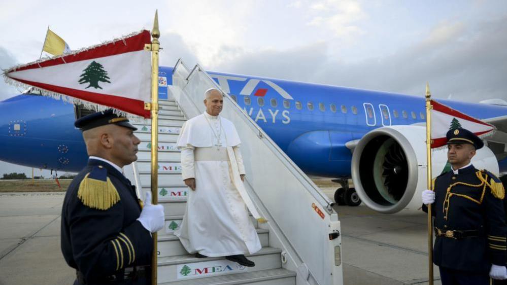 A man in white religious robes and skullcap descends the steps of an airliner as soldiers in dress uniform stand guard with flags. 