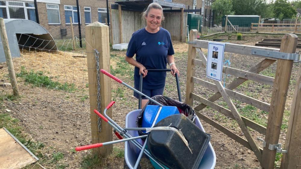Andrea Foster, holding a wheelbarrow, full of tools, by a fence, she is wearing blue shorts and a blue top. She has long hair tied back.