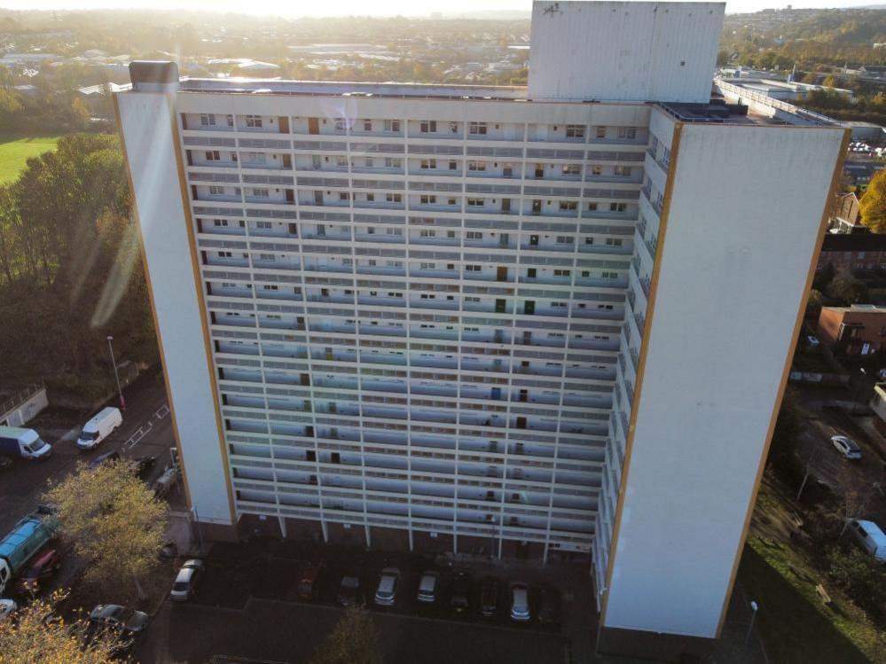 A tall white building with cars parked in front of it stands next to roads and to the left is bright green grass.