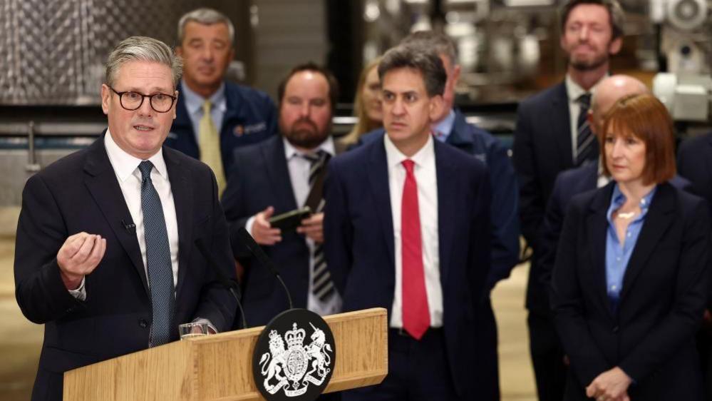 Sir Keir Starmer giving a speech next to the government podium with Rachel Reeves and Ed Miliband standing in the background