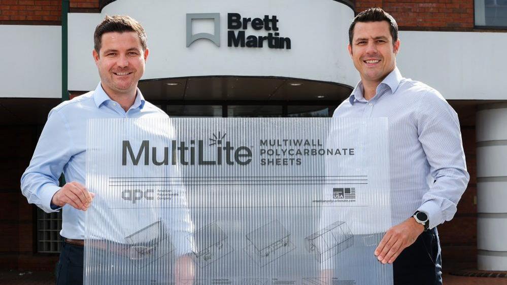 Two dark haired men in blue shirts, stood in front of a white building that says Brett Martin, they are jointly holding a translucent plastic sheet that says MultiLite Multiwall polycarbonate sheets