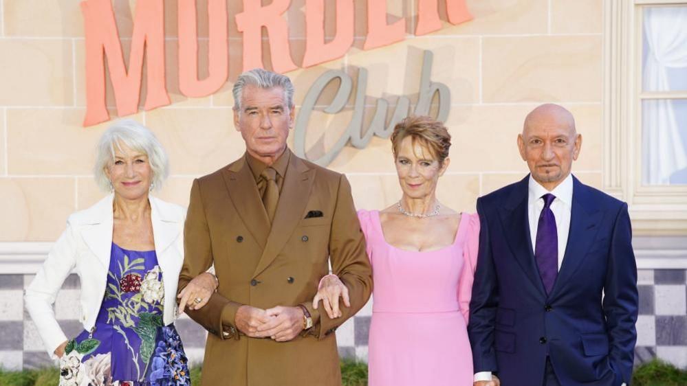 Helen Mirren in a blue dress and white jacket, Pierce Brosnan in a brown suit, shirt and tie, Celia Imrie in a pink dress  and Sir Ben Kingsley in a blue suit, white shirt and purple tie. All at looking forward and smiling.